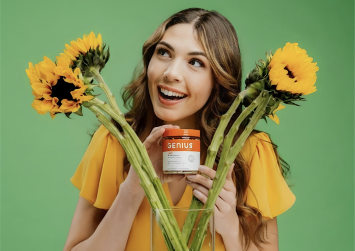 Woman smiling and holding a jar of Genius Joy supplement with sunflowers in front of her against a green backdrop.